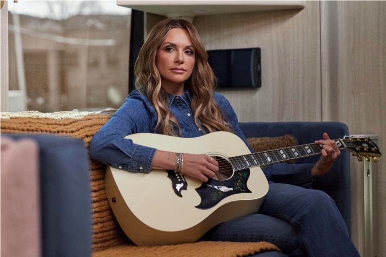 Carly Pearce holding an acoustic guitar while sitting on a couch.